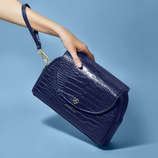 Sleek dark blue snakeskin patterned handbag held by an elegant hand on a soft blue background in lifestyle product photographer